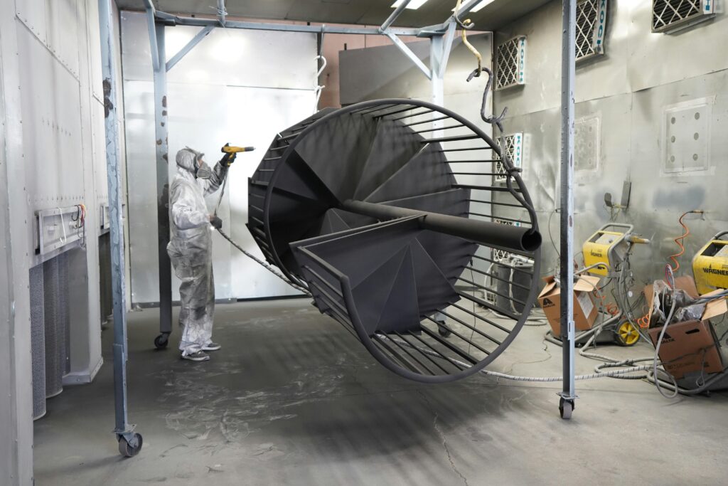 A worker in protective gear spray paints a large industrial metal structure indoors.