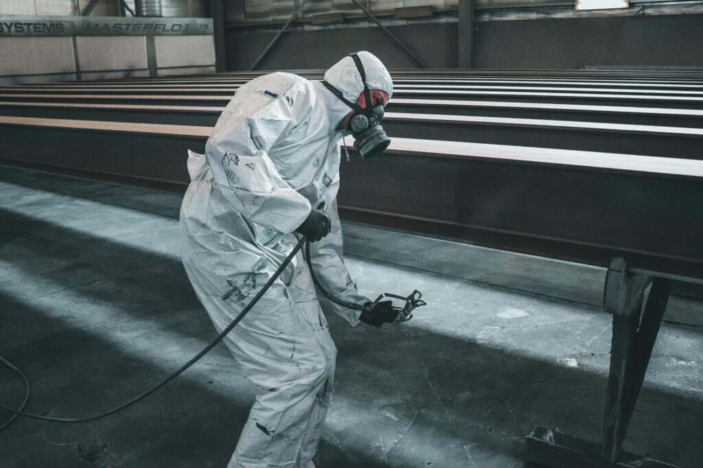 Worker in protective gear spray painting metal beams inside a factory setting.